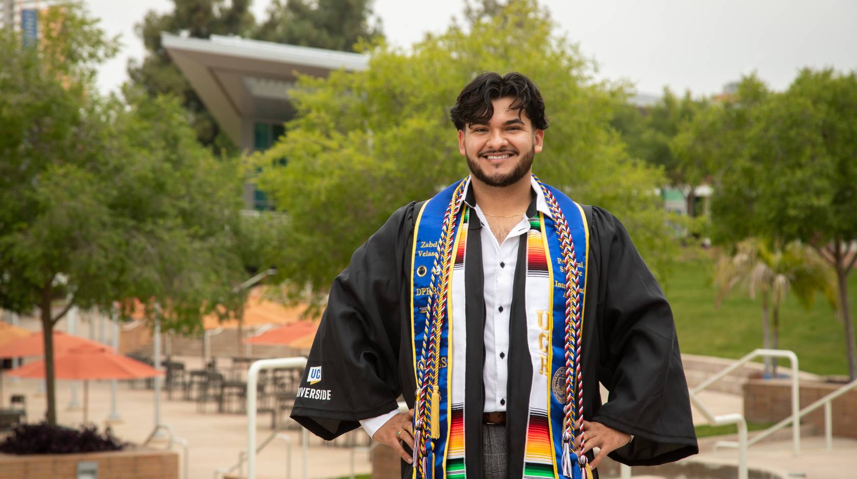 man with hands on hips wearing graduation gown and sashes on UC Riverside campus