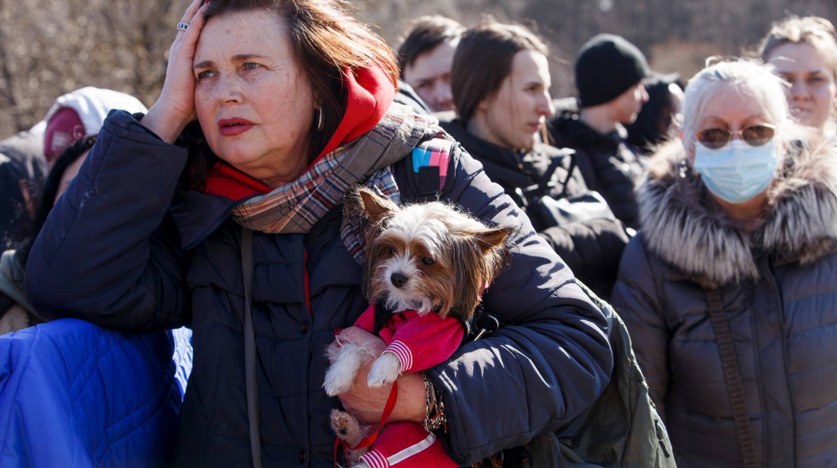 A group of Ukrainian refugees standing outside in winter coats, a middle-aged woman holding a small dog in front