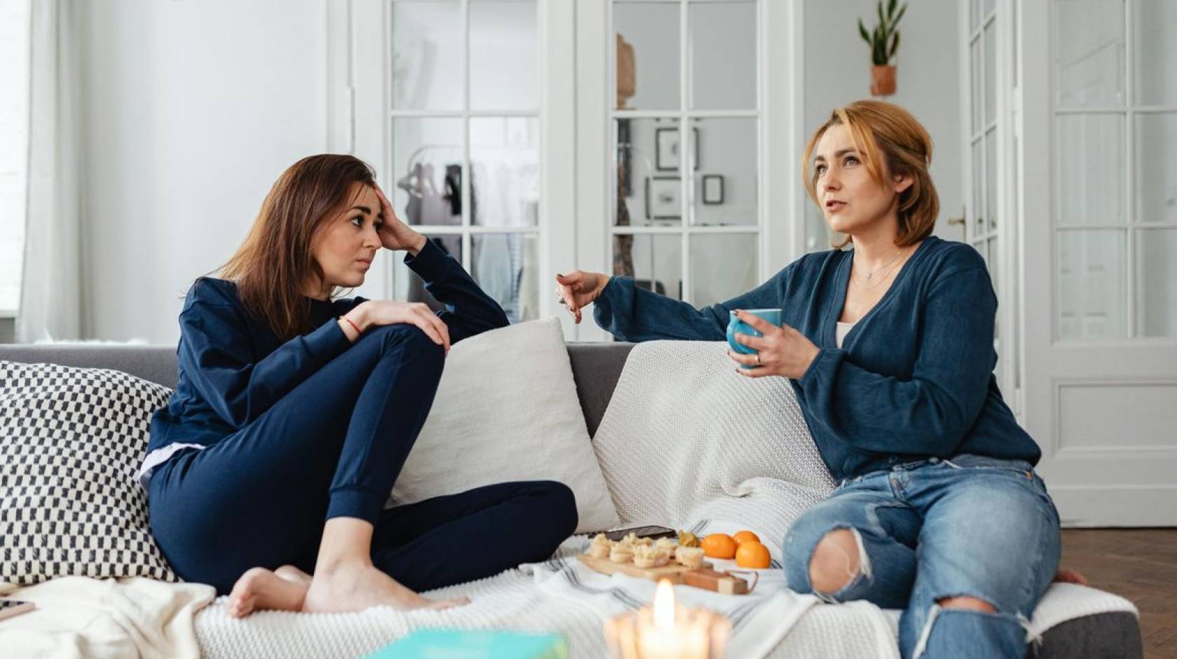 Two women sit on a couch drinking tea with a snack tray between them. The woman on the right is speaking and the woman on the left is listening.