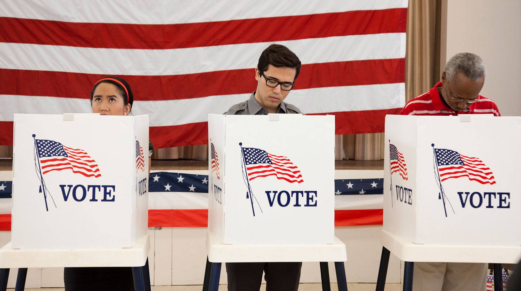 3 people stand at voting booths in front of a wall-sized American flag
