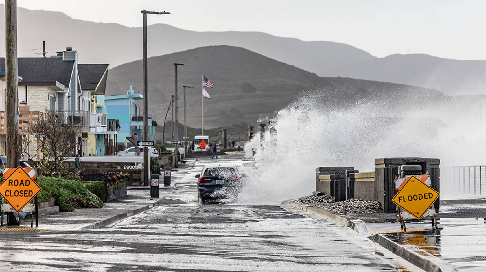 A wave crashes into a town street a blue car is driving on in Pacifica