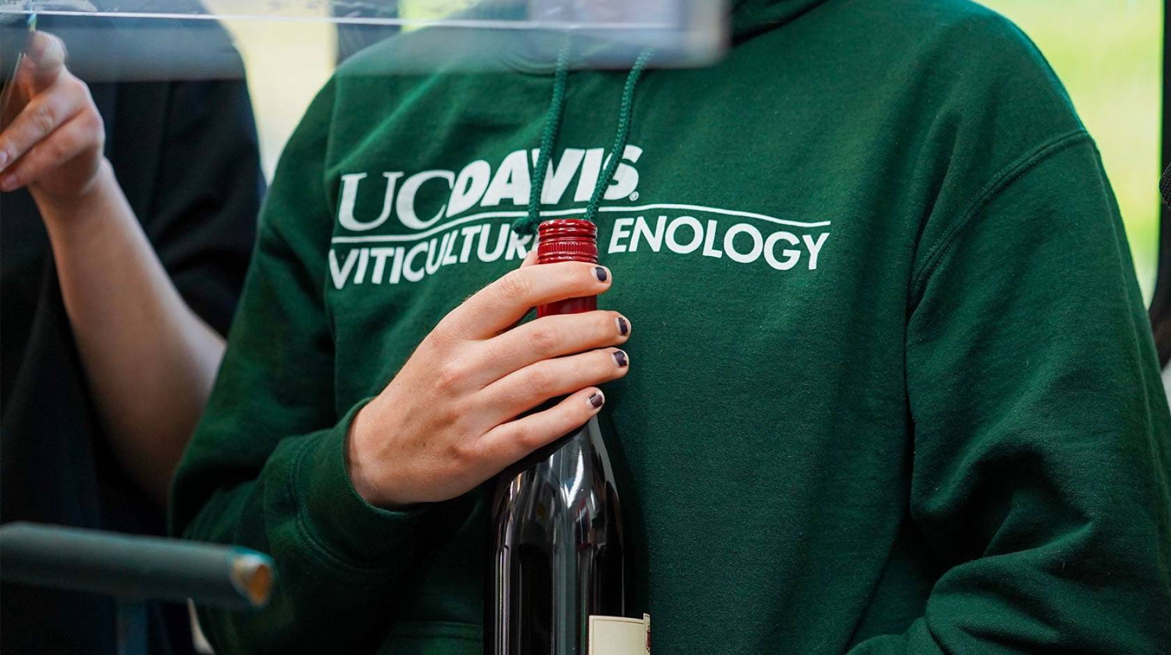 A woman in a green UC Davis viticulture sweatshirt holds a wine bottle