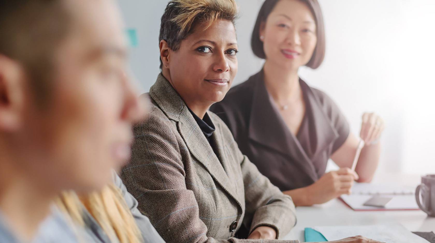 Middle-aged Black woman and middle-aged Asian woman look at a younger man in the office who is closer to the camera