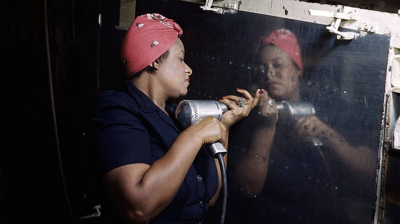 A Black woman uses a rivet gun on a reflective steel panel, wearing a red headscarf and a blue jumpsuit,