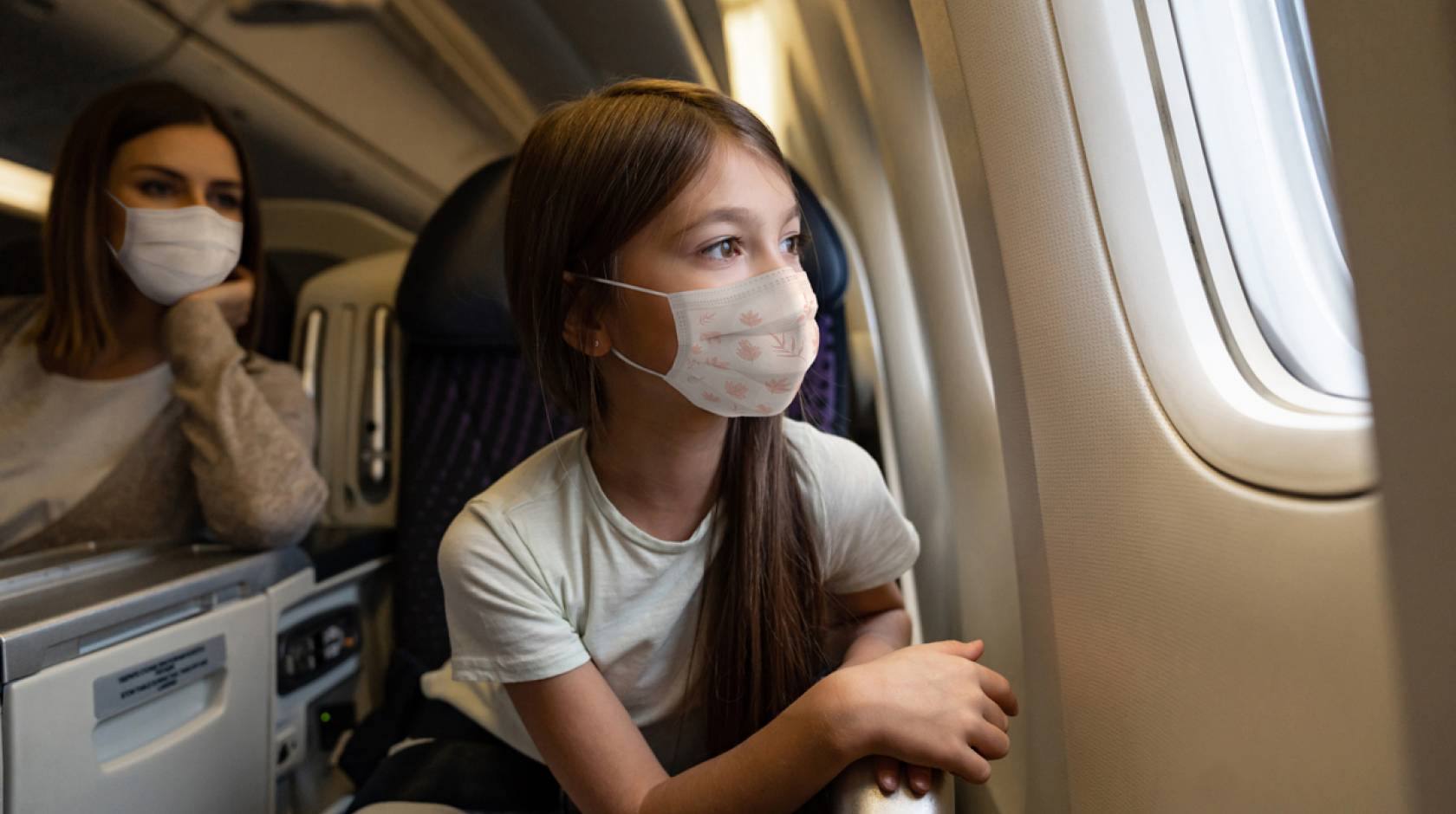 A young girl in a mask stares out an airplane window, mom in background