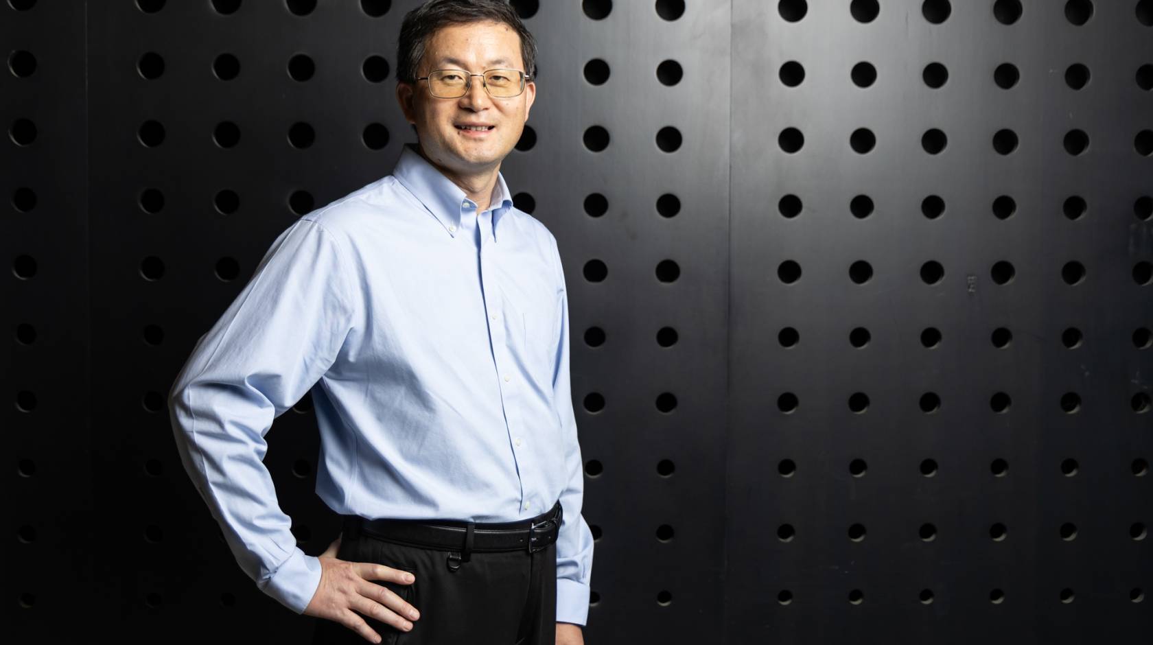 A man with glasses stands in front of a dark textured wall with his hand on his hip