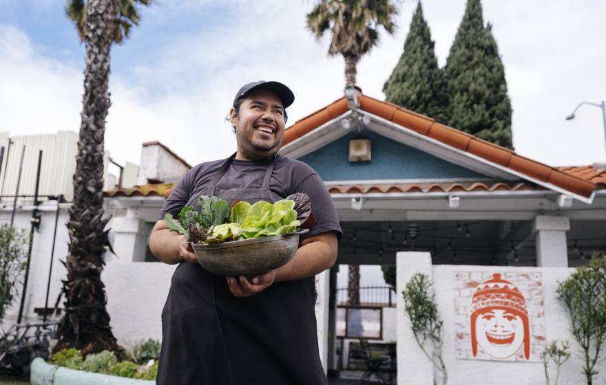 A young man smiles looking up and away from the camera, holding an armful of recently harvested lettuces. He's standing in front of a low green building with palm trees in the background. It looks like LA. 