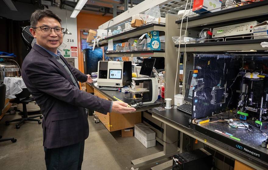 Man in a dark gray suit stands in a lab in front of multiple machines on a benchtop