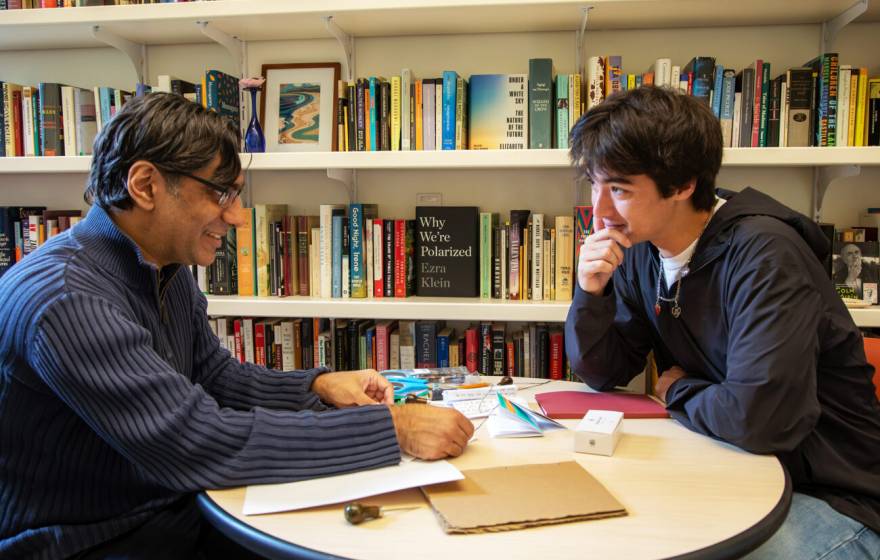A professor-aged man and a student-aged man sit at a round table in front of a bookshelf, facing each other in discussion. 
