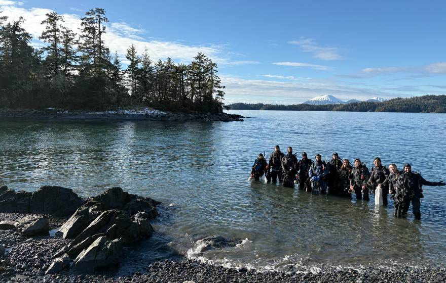A smiling group of people stands in the water in wetsuits and SCUBA gear, with evergreen trees and a snow-covered peak in the background