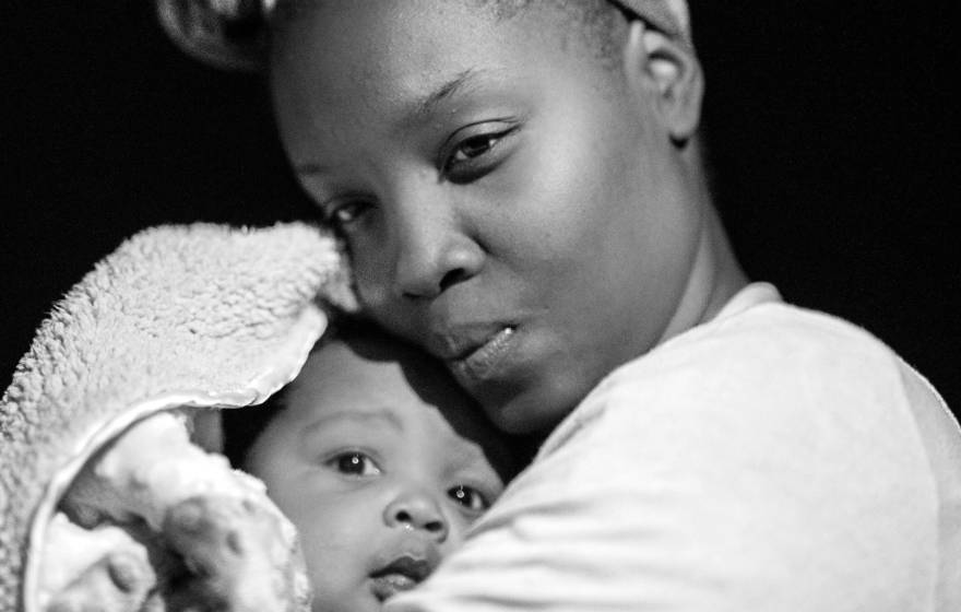 In a black-and-white close-up photo, a Black woman gazes thoughtfully as she coddles a baby wrapped in a blanket