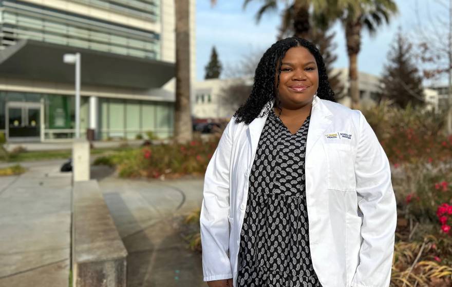 Chelsea Nash wears a black and white polka dot dress and white doctor coat, smiling for a portrait outside of a medical building at UC Davis