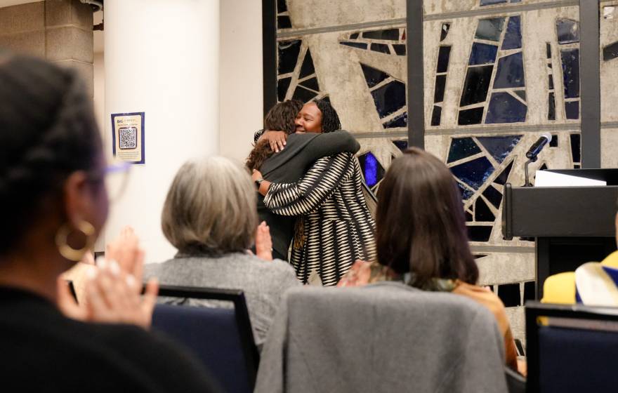 two women hug in front of a stained glass window as others applaud