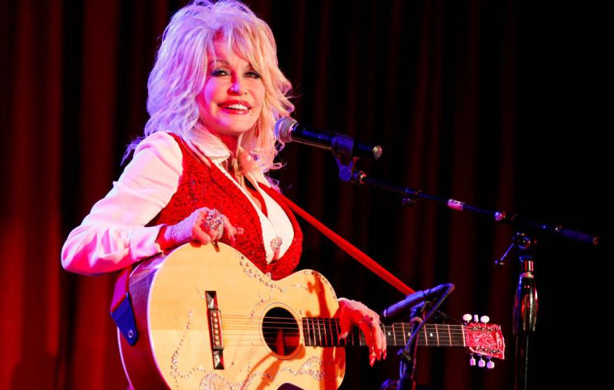 Dolly Parton in a red vest smiles while holding an acoustic guitar