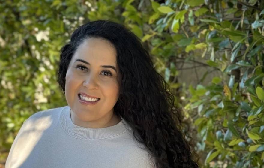 Lizbeth Lopez, 33, long curly dark hair, smiles in a shoulders-up portrait, with leaves in the background