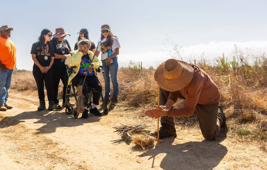 A man in a straw hat uses a wooden stick to start a fire in a brush pile while 6 people watch.