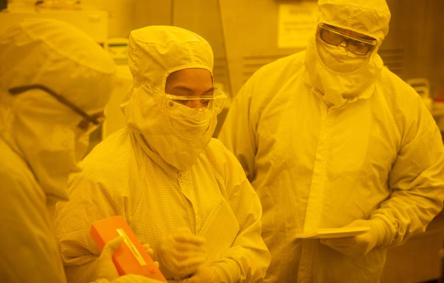 Three people stand in white full-body suits in the yellow light of Nano3's cleanroom.