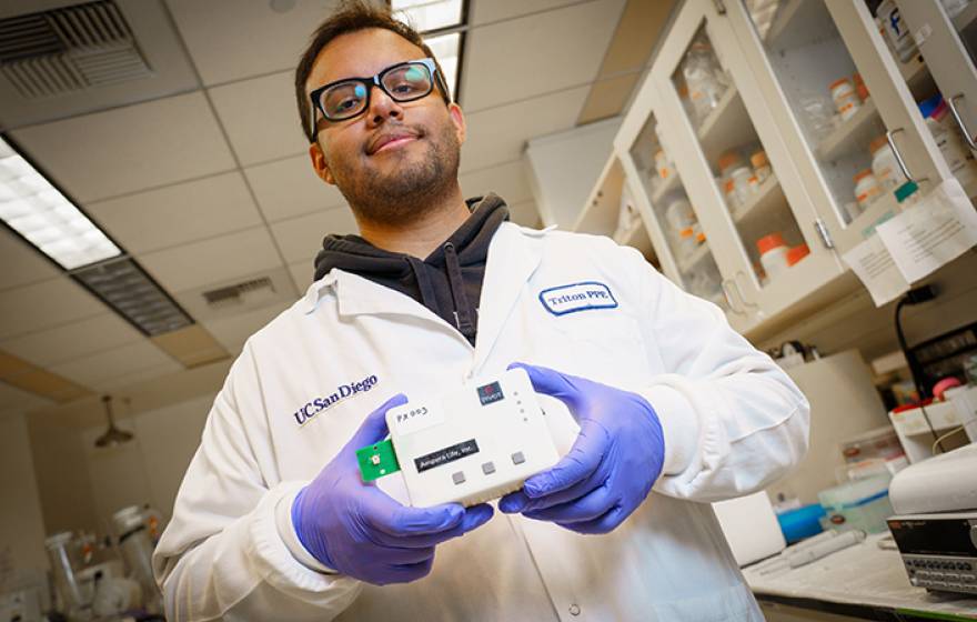 A male student in a white lab coat wearing glasses and purple rubber gloves holds a white box the size of a large sandwich in both hands