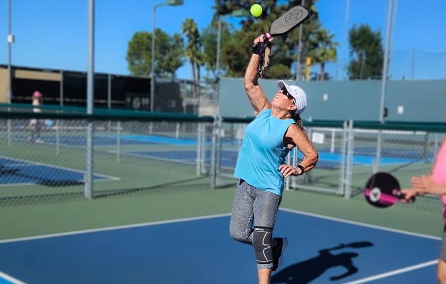 Two women on a pickleball court. A woman in the center of the frame reaches to hit a ball overhead, and her teammate looks on