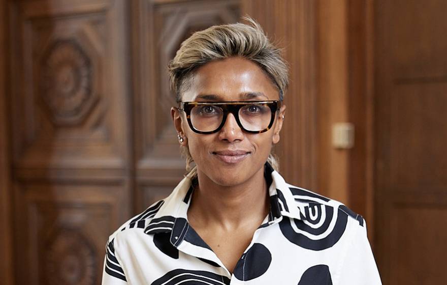 Poulumi Saha, wearing black rimmed glasses and a black and white blouse, smiles faintly for a shoulders-up portrait, with wood paneled walls in the background.