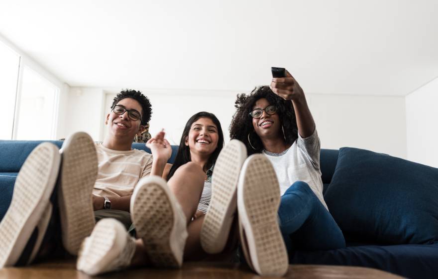 Three smiling teenagers sitting close together on a couch, one is holding a TV remote