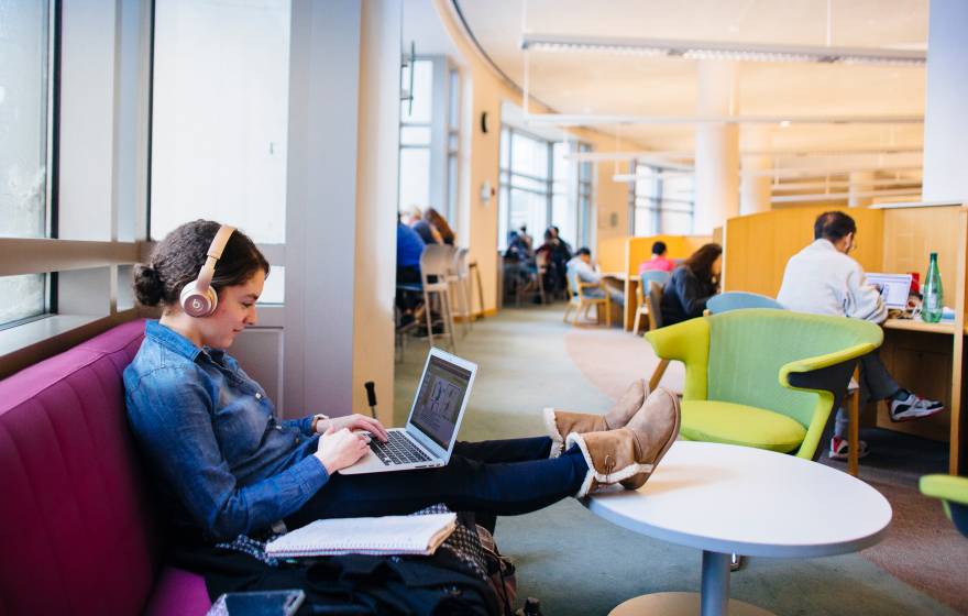 A woman sits on a sofa in a library typing on her laptop with her feet up in a table and headphones on.  Beyond her are more people studying at desks.
