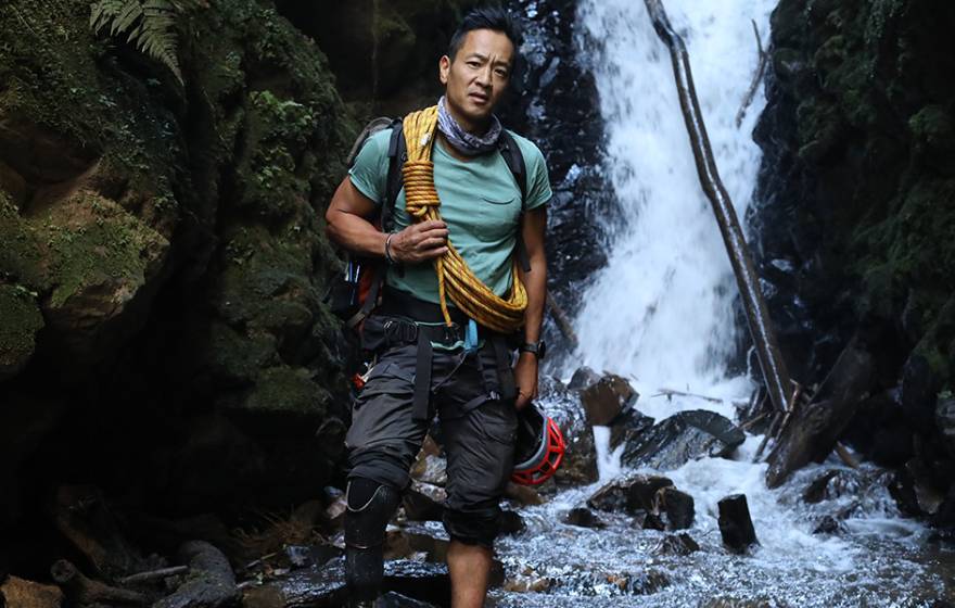 Albert Lin in outdoorsy gear stands in front of a waterfall