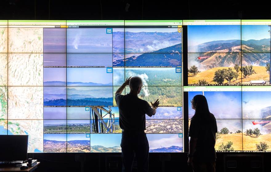 Two people in silhouette look at a large series of screens with visuals of different California landscapes, and a topographical video map of California to the left
