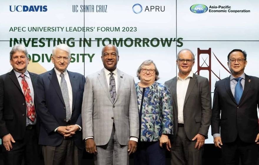 6 people in business attire stand shoulder-to-shoulder and smile for the camera in front of a video screen background reading "APEC University Leaders Forum: Investing in Tomorrow's Biodiversity"