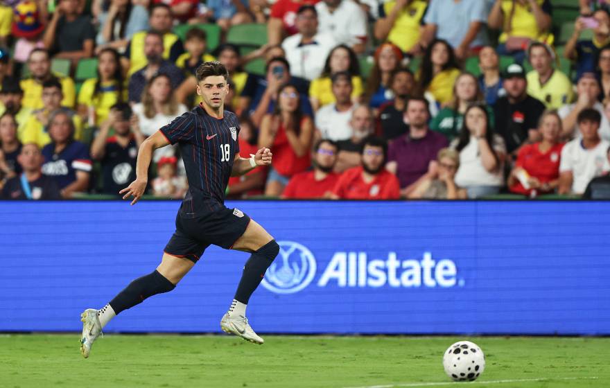 Max Arfsten of the United States in a navy blue uniform with stripes drives a ball during an international friendly match in front of the crowd, looking toward the camera