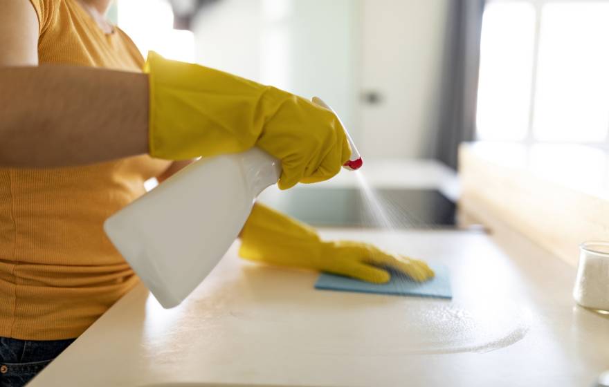 A woman wearing a peach shirt and yellow rubber gloves uses an unmarked spray bottle to clean a kitchen counter with a rag.