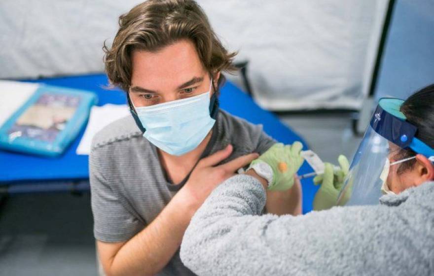 A woman wearing a face shield gives a shot to a young man wearing a face mask