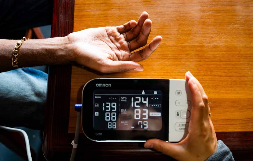 A hand is extended while another hand holds a blood pressure monitor with readings on it