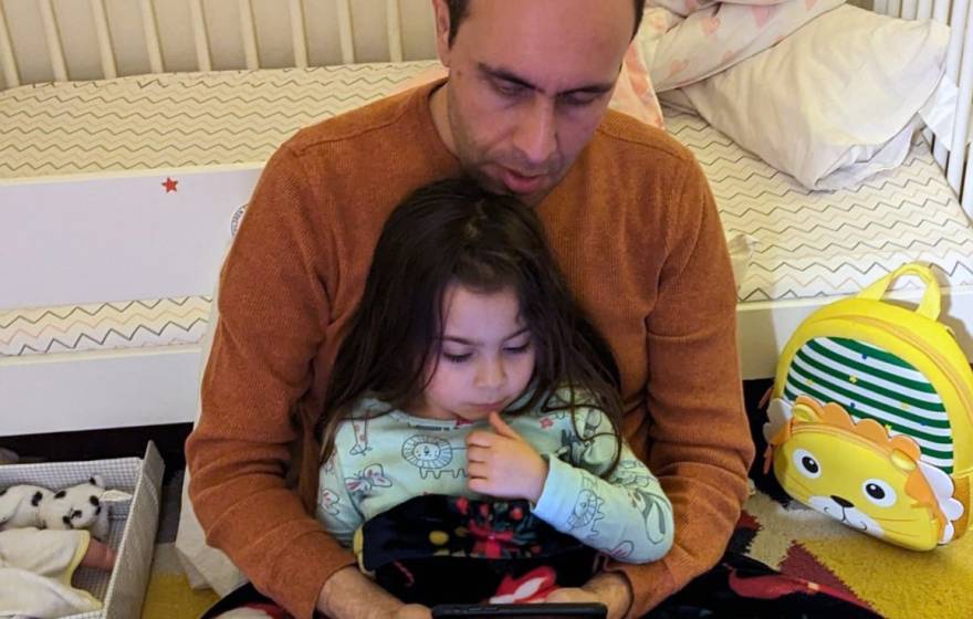 A balding man holds a young daughter as they read together, looking at a screen