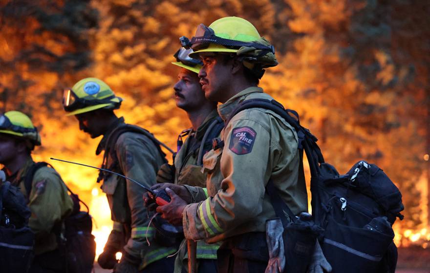 Four firefighters stand against a backdrop of flames. The scene is dark, and 2 young male firefighters hold a device with a long antenna