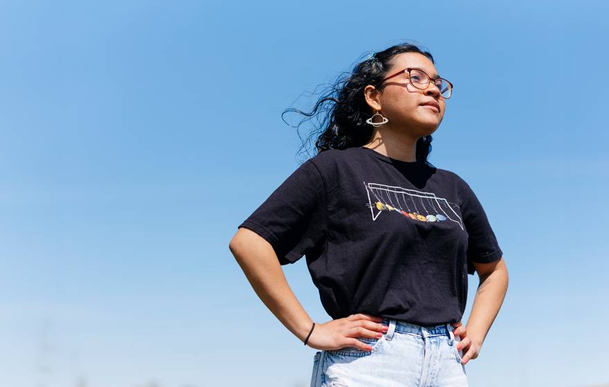 A young woman in a dark t-shirt, jeans and dangly earrings stands with her hands on her hips, looking determinedly forward, shot slightly from below, against a clear blue sky