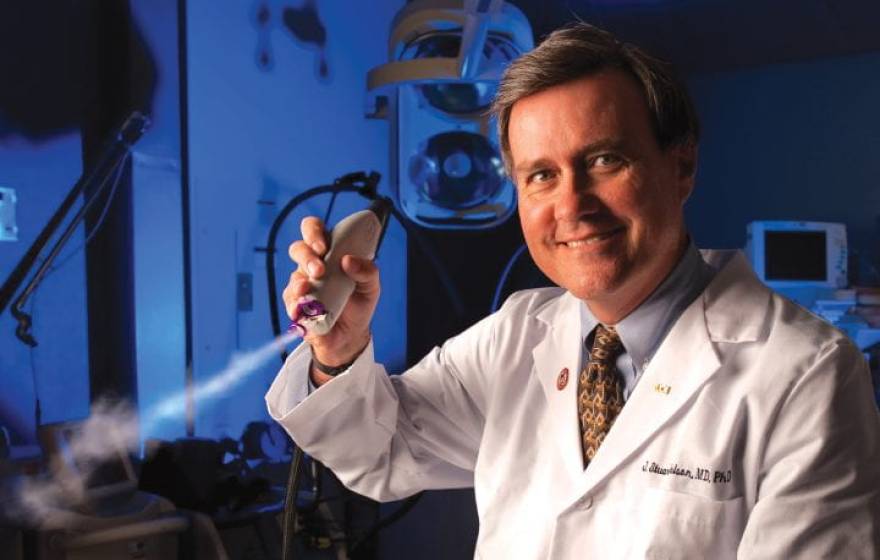 A surgeon in a white coat in a darkened lab holds a skin cooling device up to the camera and smiles.