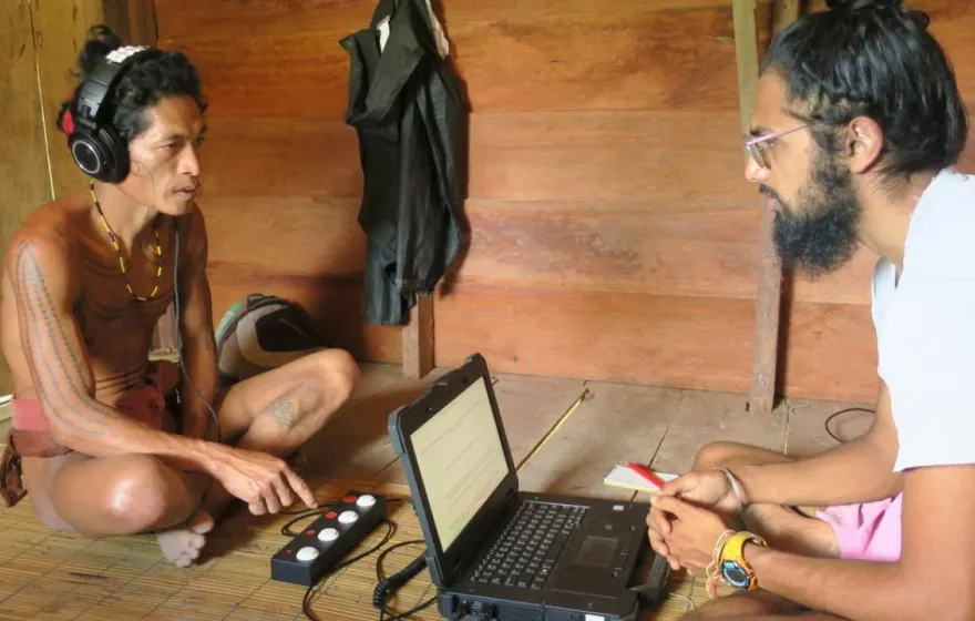 An Indigenous man sits on the floor listening to headphones, facing a researcher with a laptop