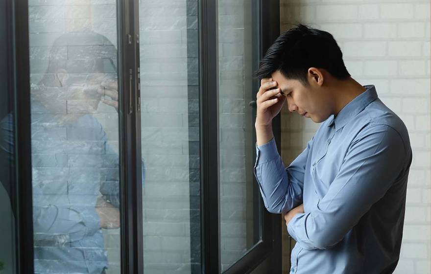 Young Asian man holds head in hand in front of his reflection at work