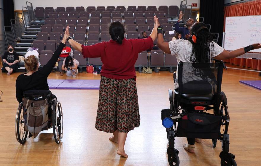 Three disabled dancers, photographed from behind, touch hands while raising their arms.