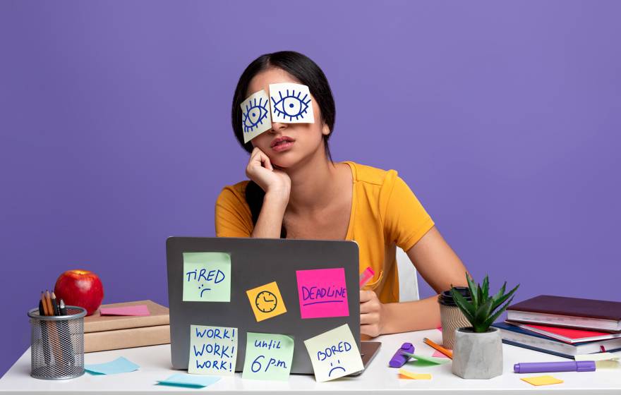 Woman with paper eyes showing her eyes open and a lot of post-its on her laptop reading Tired, Deadline, Work Work Work Until 6 p.m. and Bored