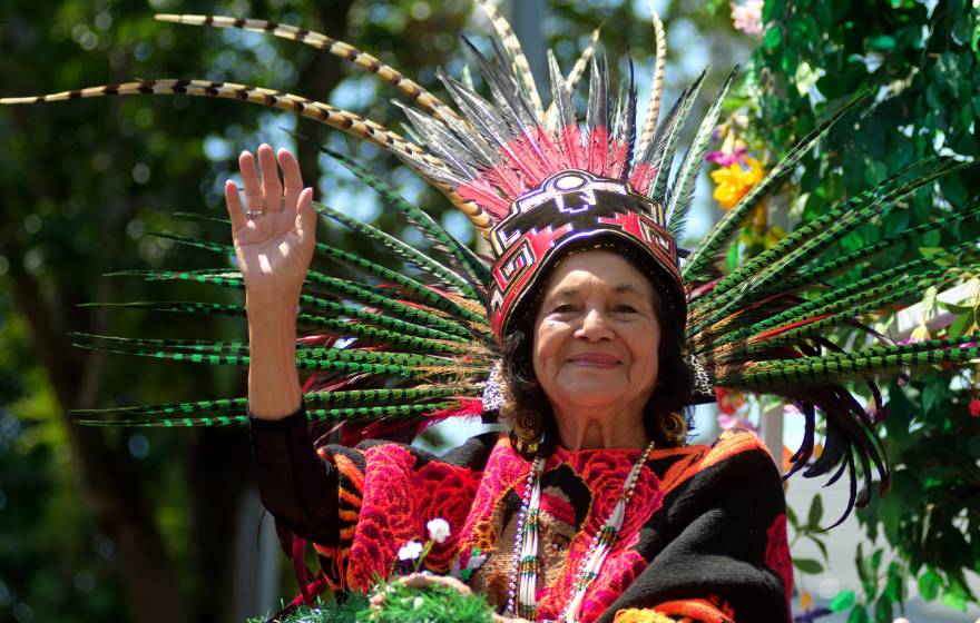 Dolores Huerta in traditional clothing and a headdress waves and smiles