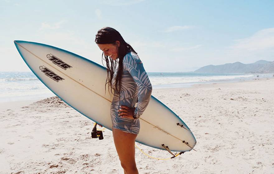 Katia Gibson on the beach with her surfboard looking down, smiling