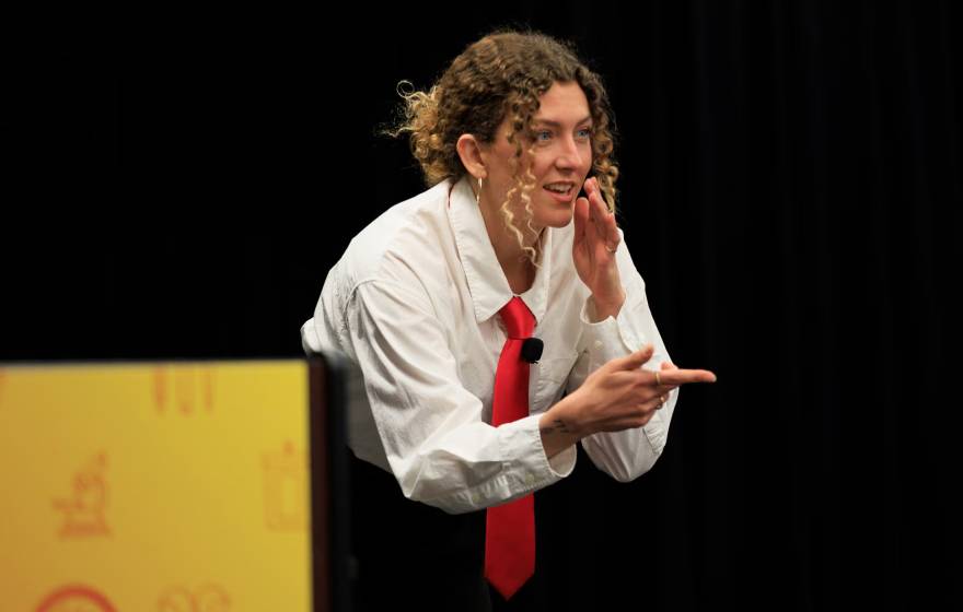 A woman in a white blouse and red tie makes a chopping gesture while pointing during a presentation