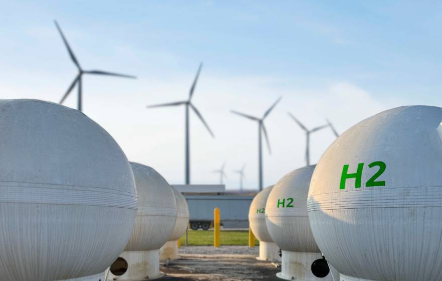 Two rows of spherical white tanks with H2 printed on them in green, with four wind turbines in the background, against a blue sky