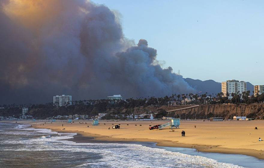 A wide sandy beach with gentle waves in the foreground, lifeguard towers and a few people scattered along the shore, while a massive dark plume of smoke rises from hills behind nearby coastal buildings under a clear blue sky.
