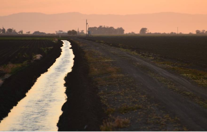 A irrigation canal runs away from the camera to a vanishing point. The fields on either side are dark, the canal reflects the orange-pink hazy sky