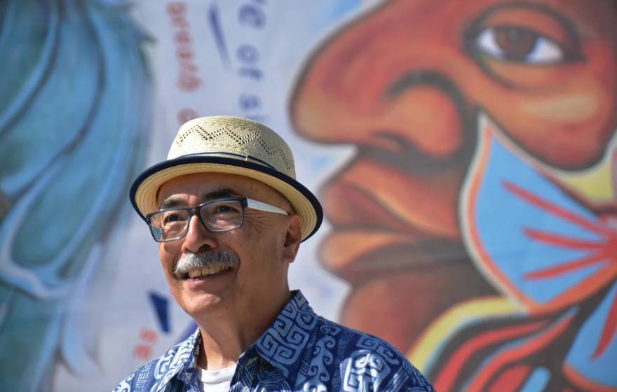 An older Latino man with a mustache, glasses and a jaunty hat in front of a colorful mural