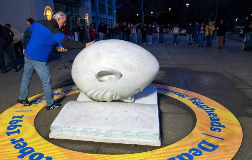 In the front of the photo, a man touches an egghead (a humorous but blank-looking human face shaped like an egg) which is faced down on a concrete open book; in the back of the photo, a line of people outside a museum at night