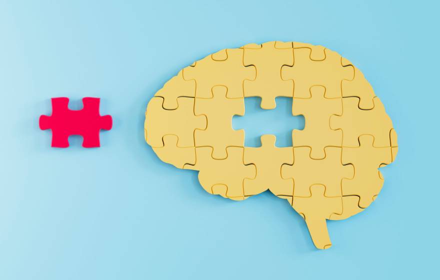 A yellow jigsaw puzzle in the shape of a brain, with one missing piece. The missing piece is red and sits beside the puzzle.